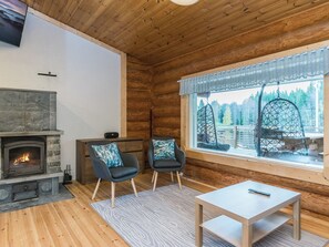 A cozy cabin interior with a stone fireplace, two armchairs, a small table, and a large window with a view of a forest.