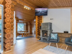Interior de una cabaña de troncos con chimenea de piedra, bancos de madera y un televisor montado en la pared.