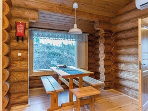 Interior de una cabaña de madera con una mesa y bancos, una ventana con vistas a árboles, y una lámpara colgante.