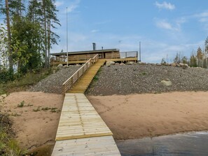A wooden boardwalk leading to a building with a sloped roof and a chimney, surrounded by trees and a rocky landscape. - Maitoniemi by Interhome (Kouvola)