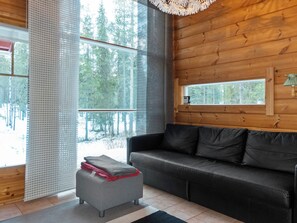 Interior acolhedor de cabana com um sofá cinza escuro, um pequeno banquinho com almofada vermelha e uma janela com vista para uma floresta.