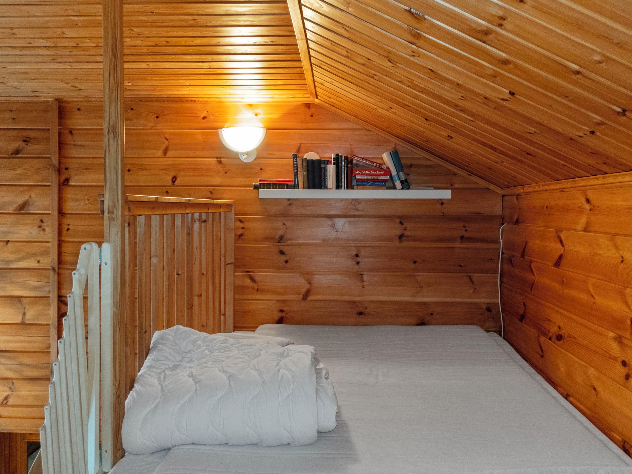 Ein kleiner, gemütlicher Raum mit Holzwänden, einer schrägen Decke, einem Einzelbett mit weißer Bettwäsche und einem Regal mit Büchern und einer Lampe.