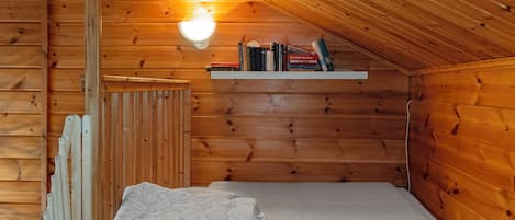 A small, cozy room with wooden walls, a sloped ceiling, a single bed with white bedding, and a shelf with books and a lamp.