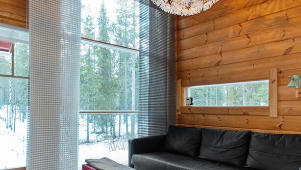A cozy room with a wooden wall, a grey ottoman, a black sofa, and a window with a view of trees. - Aisakello by Interhome (Kittilä)