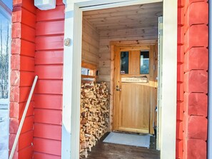 En træhytteindgang med en dør, et brændestabel og en metalringeklokke.