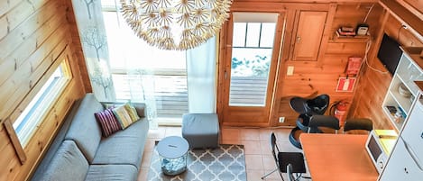 A cozy cabin interior with a sofa, ottoman, dining table, chairs, a hanging light fixture, and a kitchen area.