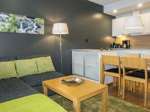 A compact living space with a sofa, a small table, a dining area with chairs, a kitchen area with a stove and cabinets, and a wall-mounted lamp. - Levi chalets 1602 by Interhome (Kittilä)