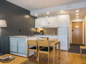 A modern kitchen with white cabinets, a built-in oven, and a coffee machine. There is a dining area with a wooden table and chairs.