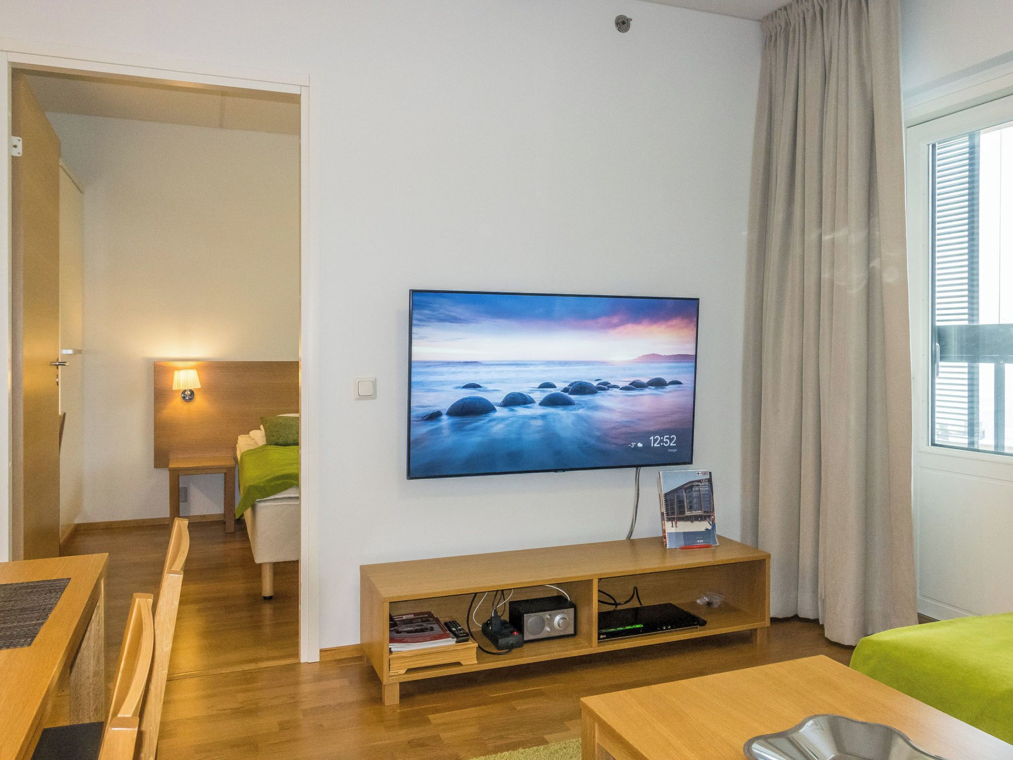 A modern hotel room with a flat-screen TV mounted on the wall, a wooden TV stand with electronic devices, a green sofa, a wooden dining table, and a view of the sea through the window.