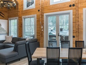 Ein modernes Interieur mit einer Holzbalkenwand, einem grauen Sofa, einem Esstisch mit Stühlen und großen Fenstern.