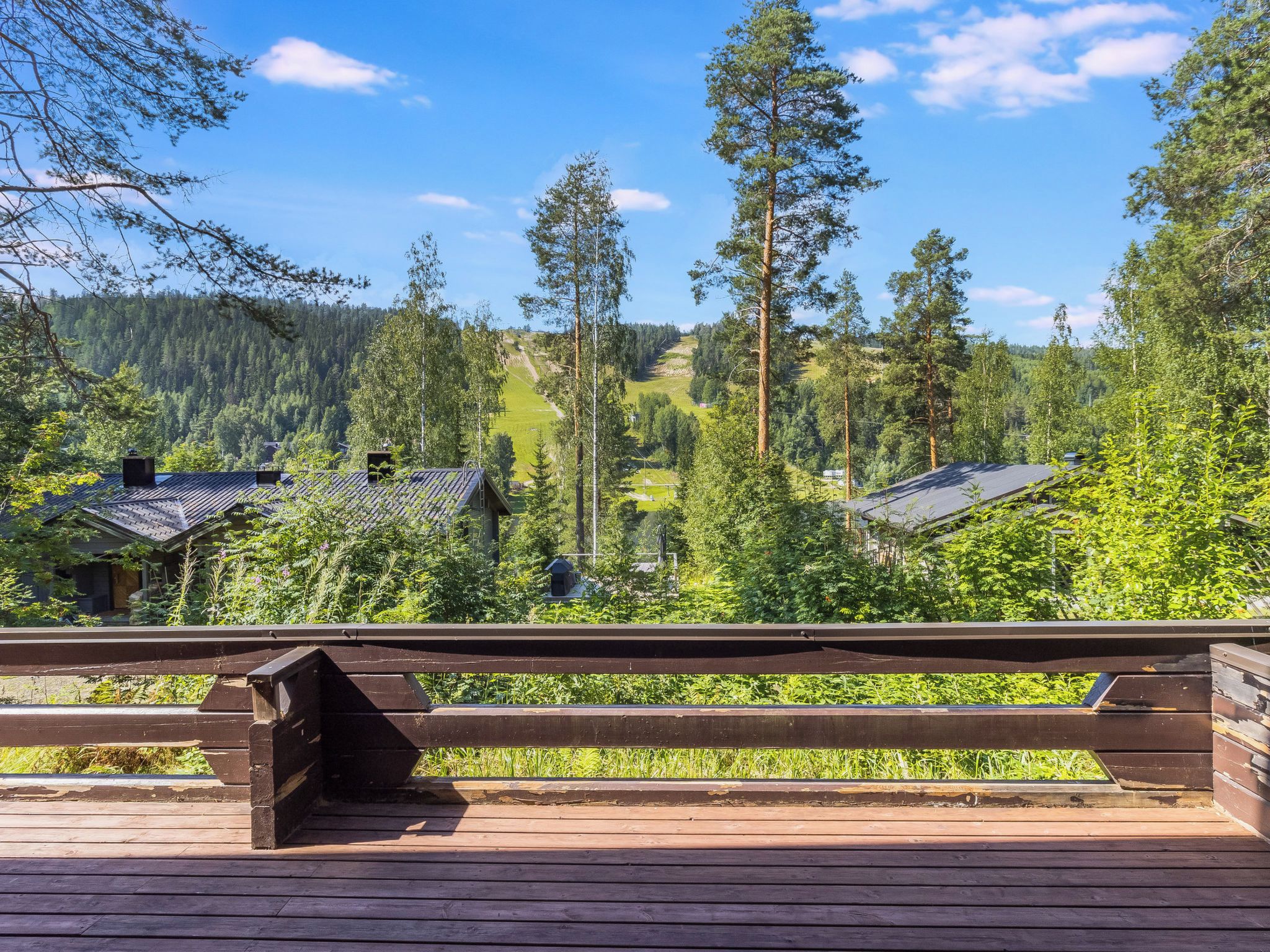 A wooden deck with a view of a forested valley, including houses with sloped roofs.