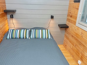 Een kleine, compacte kamer met een bed, twee kussens, een raam en een wandplank.