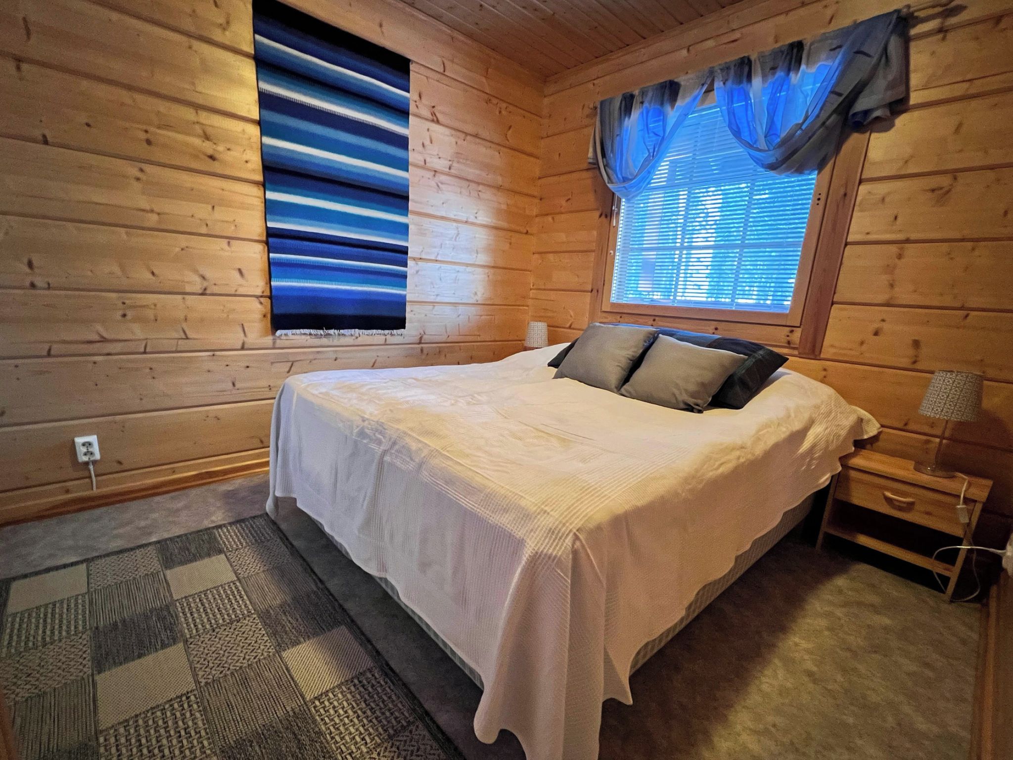 Une chambre à coucher dans un chalet en bois, avec un lit, des oreillers, une table de chevet, une lampe, une fenêtre avec des rideaux et un tapis à motifs.