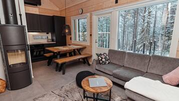 A cozy living room with a fireplace, a dining area, and large windows offering a view of a snowy forest.