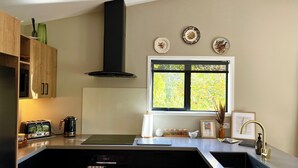 A modern kitchen with a built-in oven, a stovetop, and a black range hood. There is a window with a view of trees, and a sink with a gold faucet. - Pheasant Lodge - a touch of luxury in the countryside (Upper Moutere)