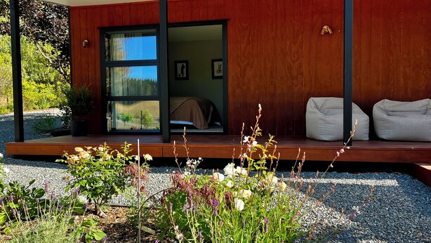 A modern outdoor seating area with a wooden deck, a glass door leading to a room with a bed and a bench, and a gravel garden with various plants. - Pheasant Lodge - a touch of luxury in the countryside (Upper Moutere)
