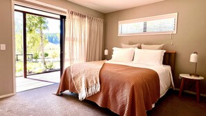 A bedroom with a large bed, a window with blinds, a bedside table with a lamp, and a view of a garden through the door. - Pheasant Lodge - a touch of luxury in the countryside (Upper Moutere)