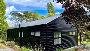A black wooden cabin with a sloped roof, surrounded by a gravel driveway and various plants. - Pheasant Lodge - a touch of luxury in the countryside (Upper Moutere)