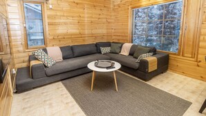 Een gezellig houten blokhutinterieur met een hoekbank, een ronde salontafel en grote ramen met uitzicht op besneeuwde bomen.