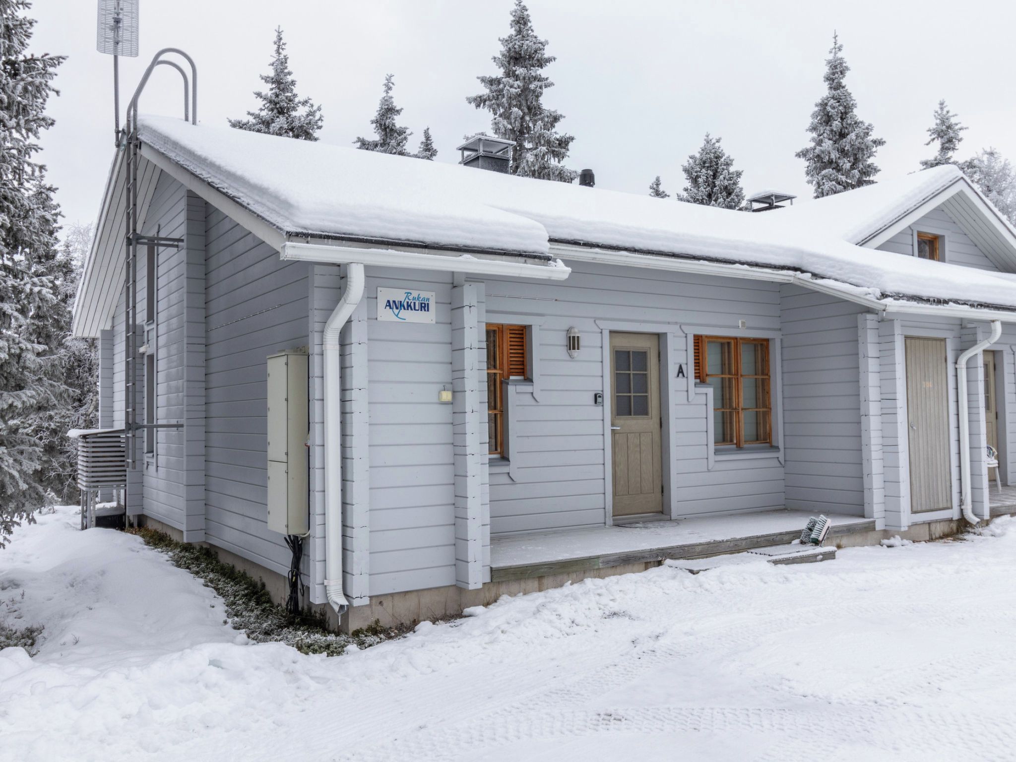 Une cabane recouverte de neige, avec une seule porte et une seule fenêtre, entourée d’arbres chargés de neige.