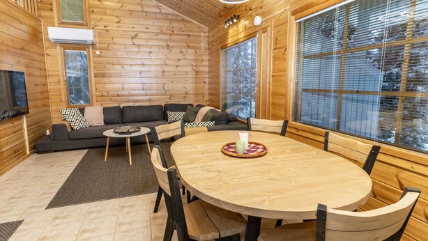 Een houten hutinterieur met een ronde eettafel, zwarte stoelen, een bank en grote ramen.