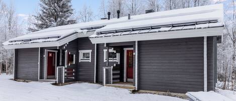Une cabane à deux étages, dont le toit est recouvert de neige, avec une porte rouge et un petit porche.