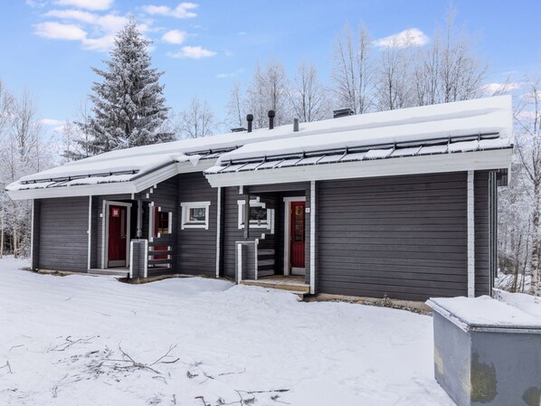 A two-story cabin with a snow-covered roof, a red door, and a small porch area.