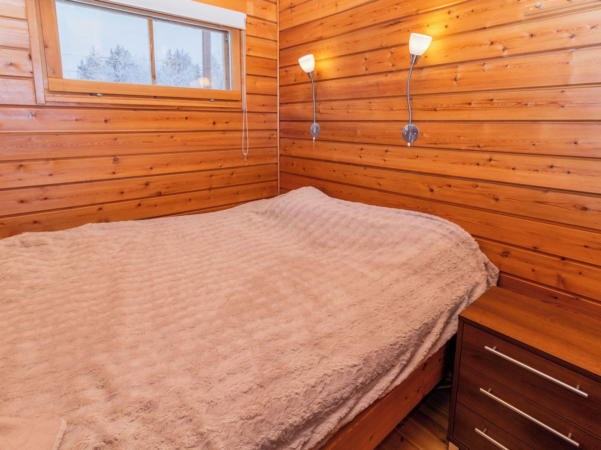 A wooden room with a bed, a window, and two wall-mounted lights.