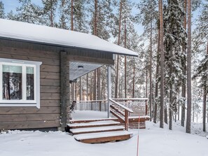 A wooden cabin with a covered porch and steps leading to a deck, surrounded by snow-covered trees.