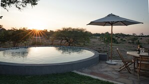 A circular pool with a stone border, surrounded by a grassy area and a wooden deck. There is a table with chairs and an umbrella, and a bottle and glass on the table.