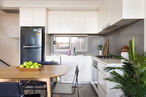 Une cuisine moderne avec des meubles blancs, un réfrigérateur en acier inoxydable, une table à manger en bois sur laquelle se trouve un saladier de pommes vertes, et une plante en pot.