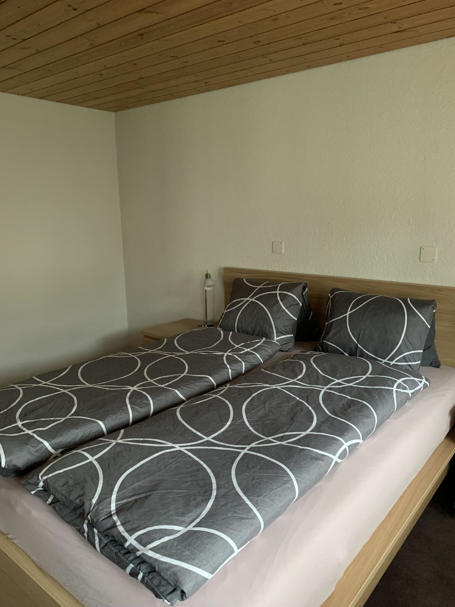 Ein Einzelbett mit grau-weißer Bettdecke, einem Holzkopfbrett und einer Nachttischlampe.