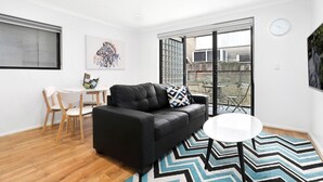 Un salon moderne avec un canapé en cuir noir, une table basse ronde blanche et un tapis à motifs chevrons. On y trouve également un coin repas avec une table et des chaises blanches, ainsi qu’une grande fenêtre donnant sur le jardin.