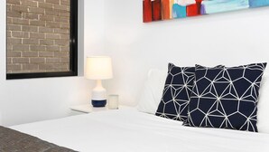 Un lit bien fait, avec une literie blanche, un coussin à motifs géométriques, une lampe de chevet et un mur en briques.