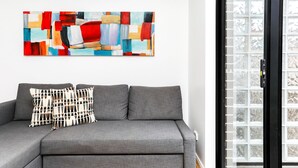 Ein graues Sofa mit zwei gemusterten Kissen, darüber ein farbenfrohes abstraktes Gemälde und ein Fenster mit Gittermuster.