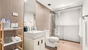 A modern bathroom with a sink, toilet, and glass-enclosed shower. There are shelves with toiletries and towels.
