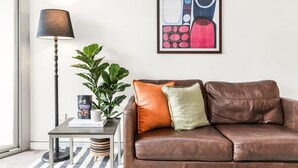 A brown leather sofa with two cushions, a floor lamp, a framed abstract painting, a small table with a book and a plant, and a window with a view.
