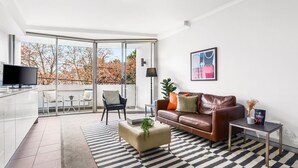 A modern living room with a sofa, coffee table, and a TV. There's a balcony with a view of trees.