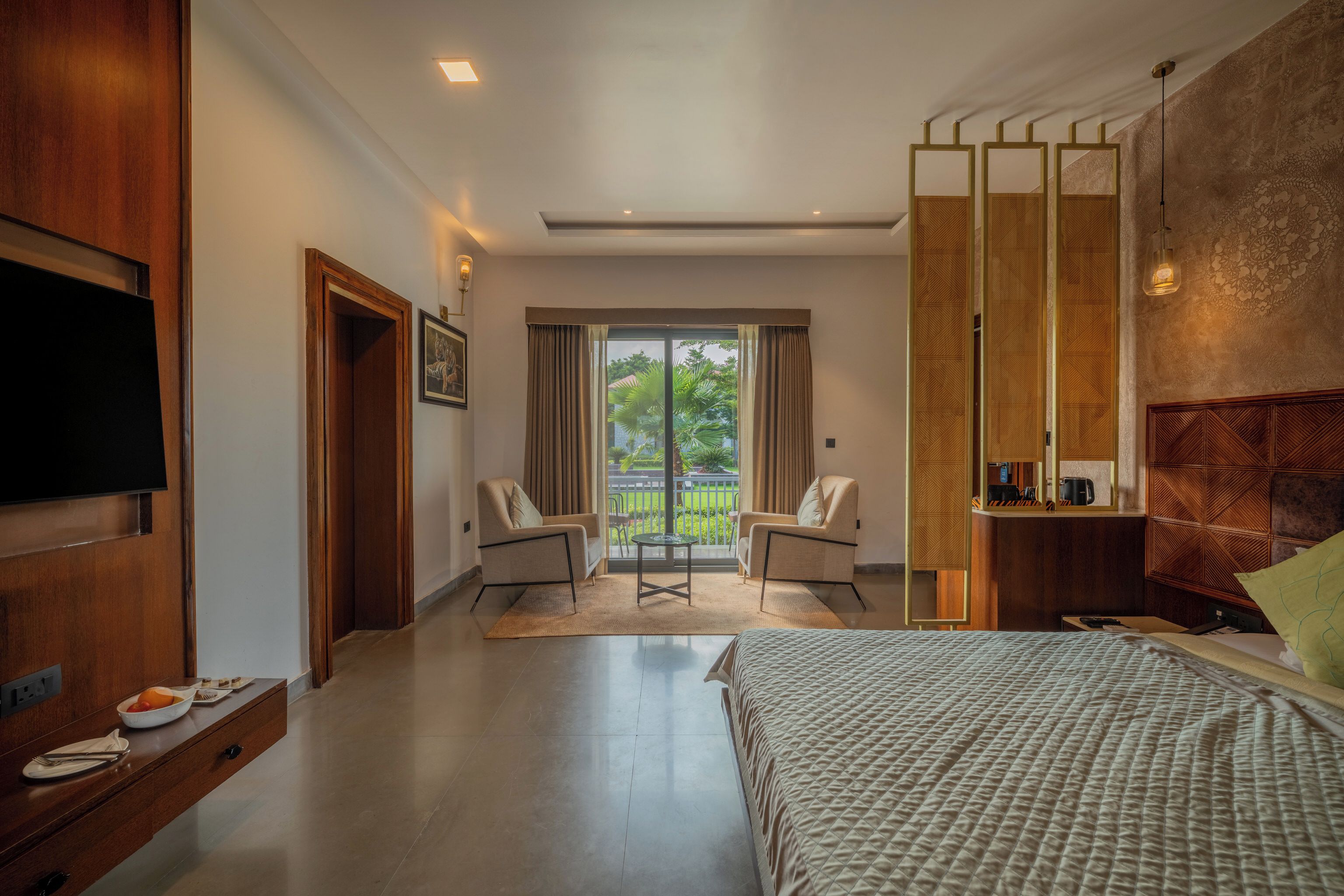 A hotel room with a bed, a TV, a seating area with chairs, a balcony with curtains, and a wooden partition.