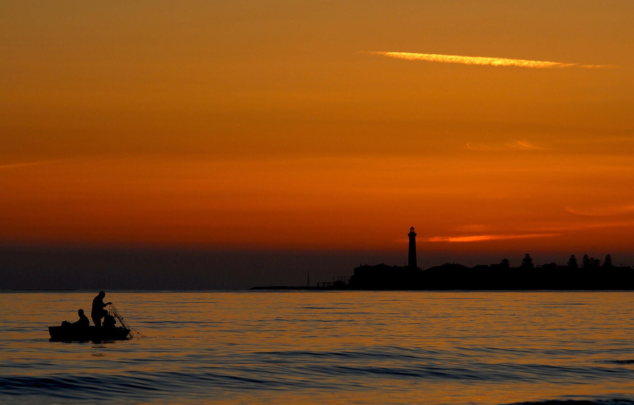 A sunset over a body of water with a silhouette of a lighthouse and a boat with people.