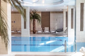 Zona de piscina interior moderna con agua azul cristalina, sillas de descanso y una palmera.