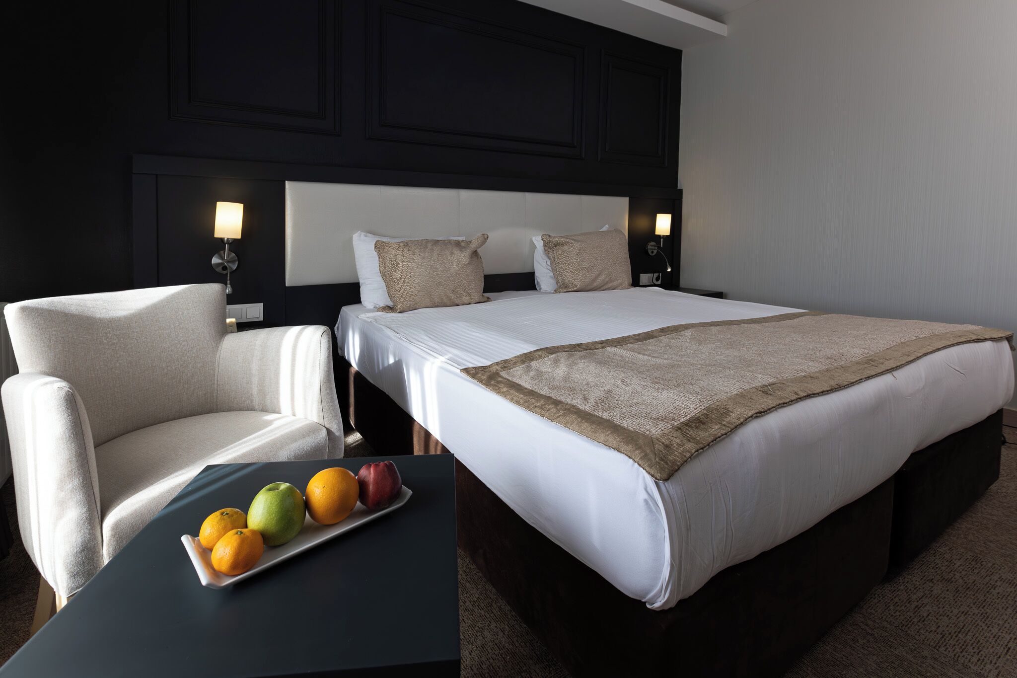 A hotel room with a large bed, two bedside lamps, a chair, and a table with fruit.
