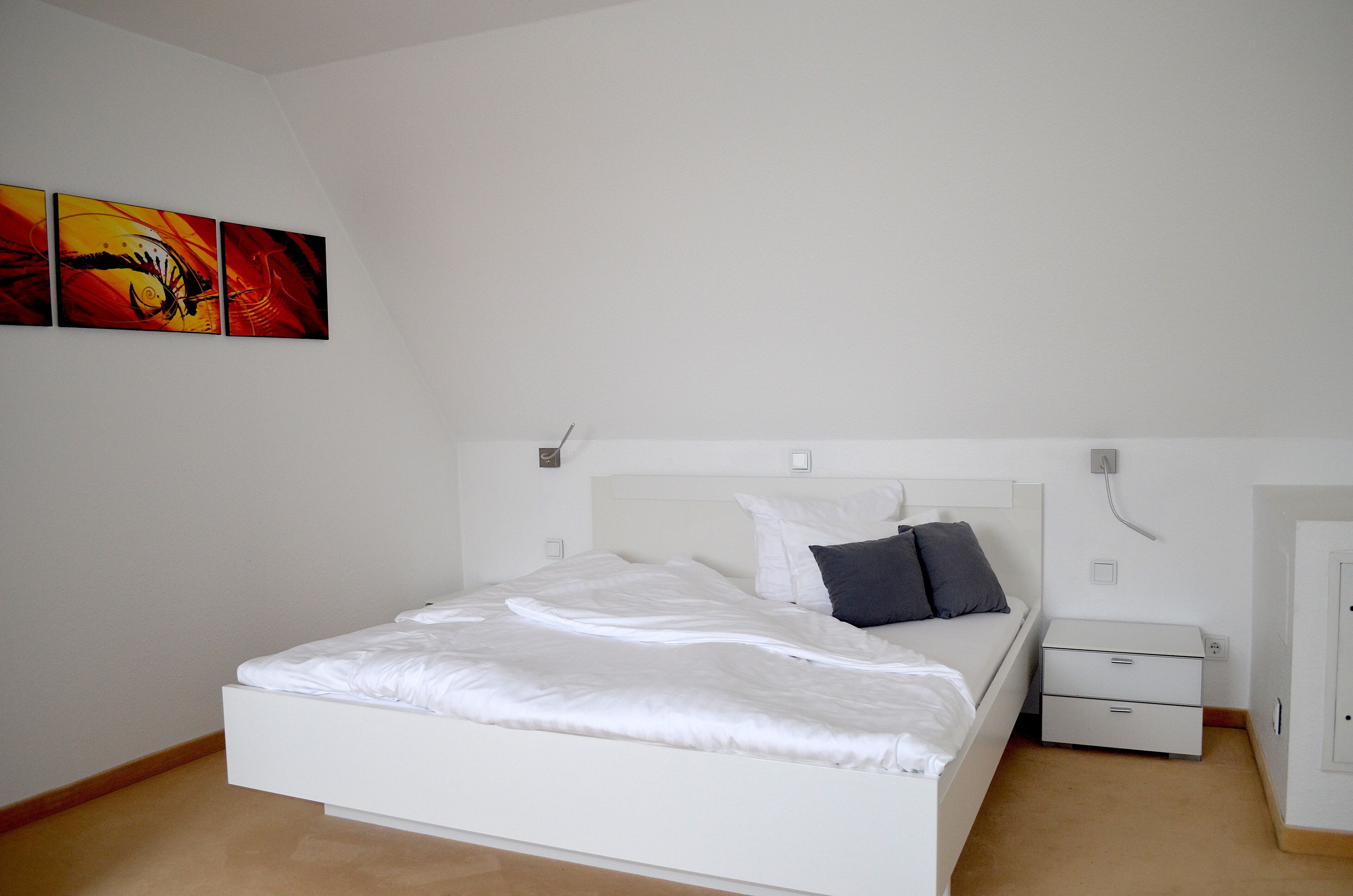 A bedroom with a white bed, a nightstand, a dresser, and three paintings on the wall.