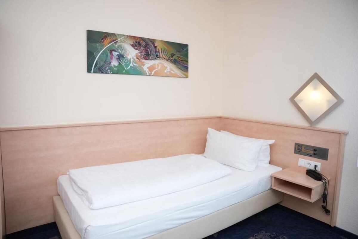 Quarto de hotel com uma cama de solteiro, mesa de cabeceira com telefone e luminária fixada na parede.