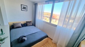 Une chambre à coucher avec un lit, une fenêtre avec des rideaux, une citation encadrée au mur et une vue sur l’extérieur.