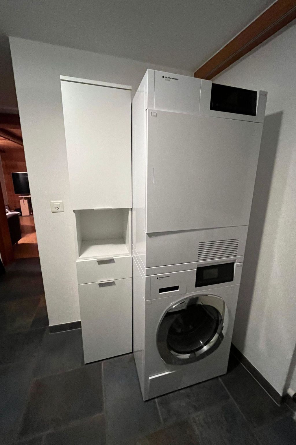 Ein kompaktes, weißes, wandmontiertes Wasch- und Trocknergerät mit einem kleinen Stauraumschrank und einer Bedienkonsole.