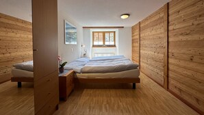 A bedroom with a wooden floor, a bed, a nightstand, a dresser, a window, and a ceiling light. - Ferienhaus Lomatte 1758 by Interhome (Saas-Fee)