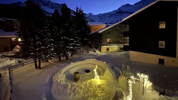 A snow-covered courtyard with a wooden barrel, a tree, and a building with multiple windows. - Ferienhaus Lomatte 1758 by Interhome (Saas-Fee)