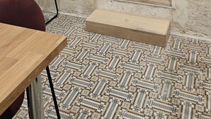 Una habitación con una mesa de madera, una silla roja, un piso estampado, una cama con cabecera de madera y una pared de ladrillo visto y enlucido.