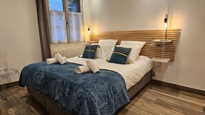 Una cama bien tendida con un edredón azul y dos almohadas, una mesita de noche con una lámpara y una ventana con cortinas.